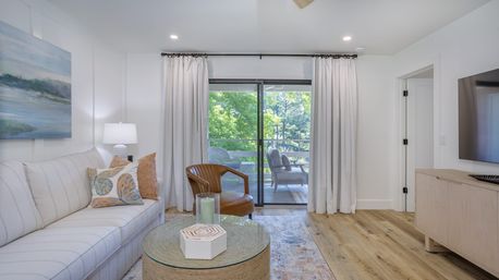 Bright modern living room with a neutral striped sofa, leather accent chair, round woven coffee table and sliding glass doors opening to a leafy balcony, hardwood floors and light curtains.