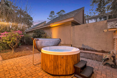Cozy backyard hot tub with wooden surround on brick paver patio at dusk, small steps, flowering shrubs and a privacy wall