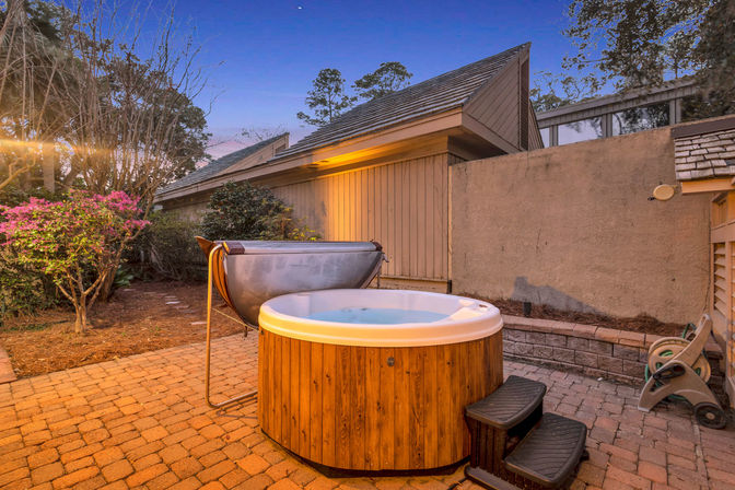 Cozy backyard hot tub with wooden surround on brick paver patio at dusk, small steps, flowering shrubs and a privacy wall