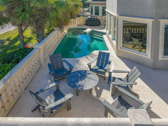 Sunlit poolside patio with six gray Adirondack chairs around a round table, balustrade railing, palm trees and a small turquoise private pool beside a modern house.