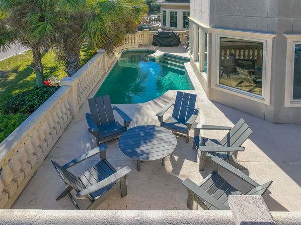 Sunlit poolside patio with six gray Adirondack chairs around a round table, balustrade railing, palm trees and a small turquoise private pool beside a modern house.