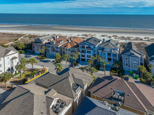 Aerial view of beachfront homes and palm trees along a wide sandy beach with calm blue ocean and horizon under a bright sky — coastal neighborhood beach houses.