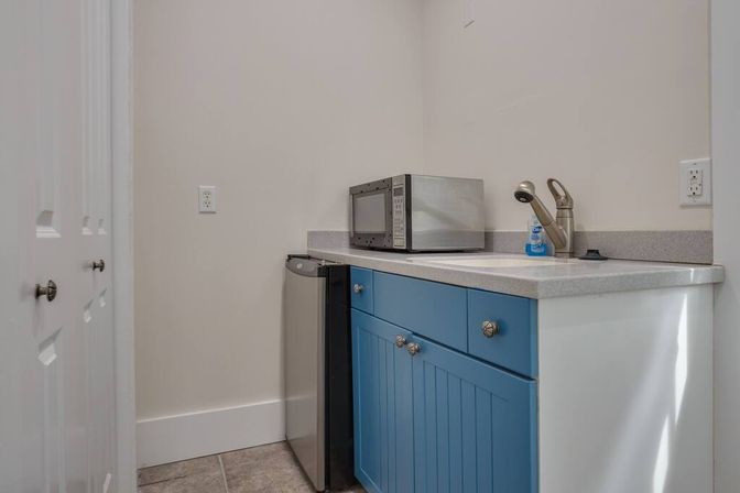 Cozy compact kitchenette with blue cabinet doors, gray countertop holding a stainless microwave, mini fridge, and sink with single-handle faucet in a neutral-walled utility corner.