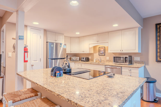 Bright open-concept residential kitchen with a large granite island, stainless-steel appliances, white cabinets, glass cooktop, microwave, and woven bar stools