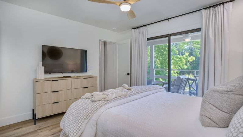 Bright modern bedroom with white textured bedding, light wood dresser and wall TV, sliding glass doors to a balcony with chairs and lush green wooded view