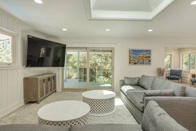 Sunlit modern living room with gray sectional sofa, two round white lattice coffee tables, wall-mounted TV, and sliding glass doors opening to a tree-lined balcony.