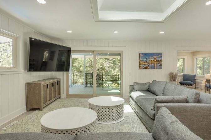 Sunlit modern living room with gray sectional sofa, two round white lattice coffee tables, wall-mounted TV, and sliding glass doors opening to a tree-lined balcony.