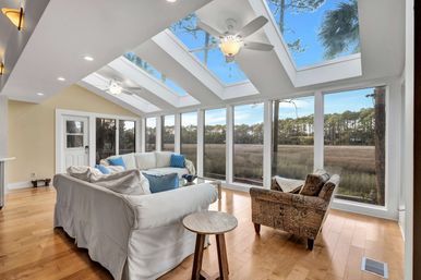 Bright sunroom with vaulted skylights and ceiling fans, white slipcovered sofas and patterned armchair on hardwood floors, floor-to-ceiling windows framing a coastal marsh and pine tree treeline.