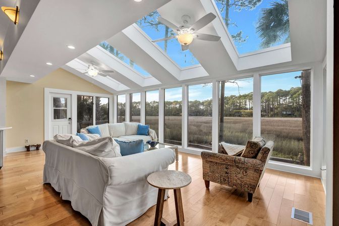 Bright sunroom with vaulted skylights and ceiling fans, white slipcovered sofas and patterned armchair on hardwood floors, floor-to-ceiling windows framing a coastal marsh and pine tree treeline.