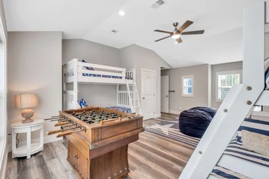 Playful coastal-style kids’ game room with white bunk beds, vintage wooden foosball table, navy bean bag, striped rug, lamp and ceiling fan in a bright beach-house interior.