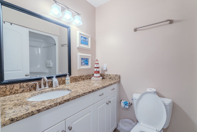 Bright coastal-themed bathroom with granite countertop vanity, undermount sink, large framed mirror, white cabinets, toilet and lighthouse decor