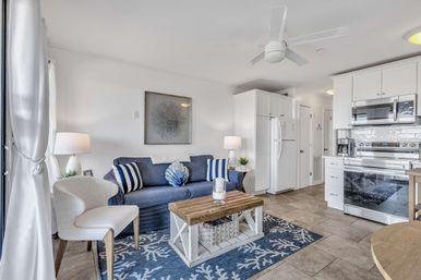 Bright open-plan coastal living room and kitchen with a navy sofa, striped and shell pillows, white armchair, rustic wood coffee table on a blue coral rug, white cabinets, stainless appliances, tile floor and ceiling fan.