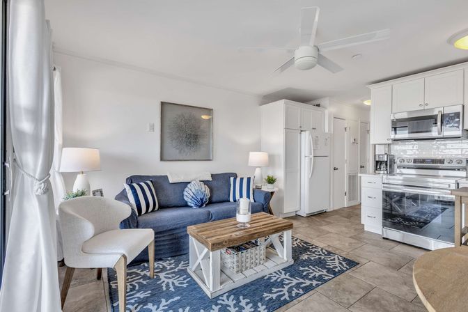 Bright open-plan coastal living room and kitchen with a navy sofa, striped and shell pillows, white armchair, rustic wood coffee table on a blue coral rug, white cabinets, stainless appliances, tile floor and ceiling fan.