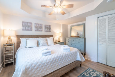 Sunlit coastal-style bedroom with wicker headboard, white quilted king bed topped with folded towels, matching bedside lamps, blue dresser with mirror and ceiling fan.