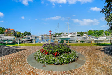 Sunny coastal marina under blue skies with moored yachts, a red-and-white striped lighthouse in the harbor, and a circular brick plaza with a vibrant flower bed in the foreground.