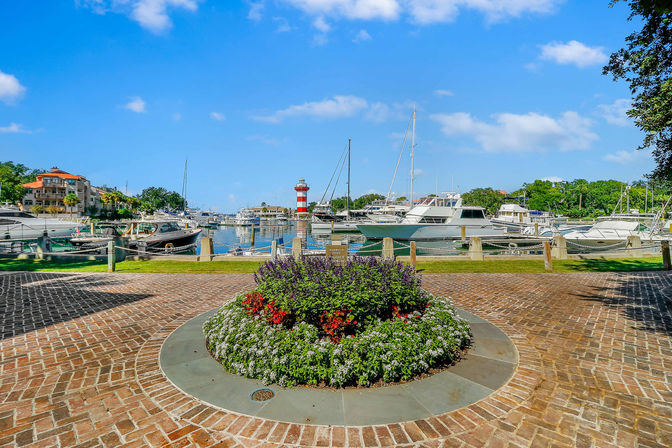 Sunny coastal marina under blue skies with moored yachts, a red-and-white striped lighthouse in the harbor, and a circular brick plaza with a vibrant flower bed in the foreground.