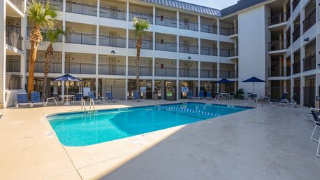 Sunny hotel courtyard with a clear blue rectangular outdoor swimming pool, lounge chairs and umbrellas, palm trees, and four-story balconies overlooking the water.