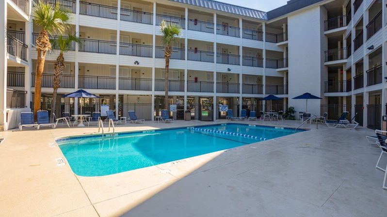 Sunny hotel courtyard with a clear blue rectangular outdoor swimming pool, lounge chairs and umbrellas, palm trees, and four-story balconies overlooking the water.