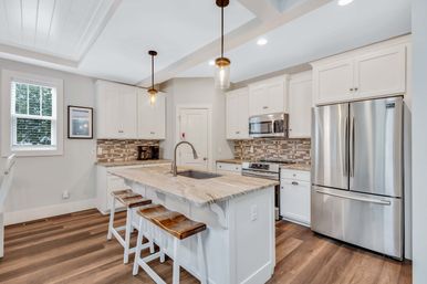 Bright modern open-concept kitchen with white cabinets, marble island countertop and undermount sink, three wooden bar stools, stainless steel French-door refrigerator and range, mosaic tile backsplash, pendant lights and warm wood flooring.