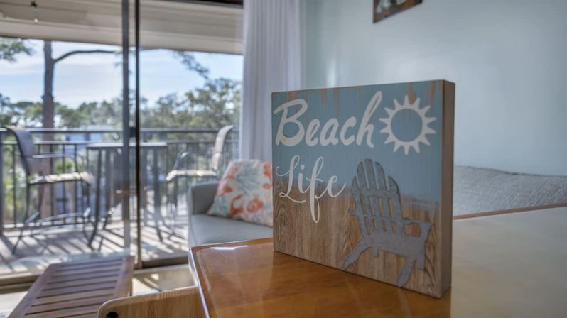 Wooden “Beach Life” sign on a table in a bright coastal condo living room, sliding glass doors opening to a balcony with chairs and a water view.