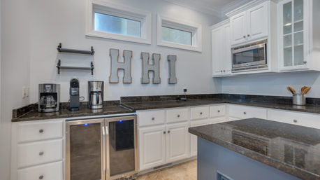 Bright modern kitchen with white cabinetry and dark granite countertops, kitchen island, built-in stainless microwave, double beverage fridge, coffee station, high windows and decorative HHI wall letters.