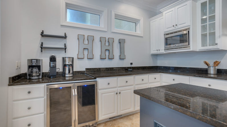 Bright modern kitchen with white cabinetry and dark granite countertops, kitchen island, built-in stainless microwave, double beverage fridge, coffee station, high windows and decorative HHI wall letters.