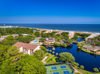 Aerial view of a sunny oceanfront resort with a central lagoon and fountain, pool and lounge areas, palm‑lined lawns, tennis courts, and the beach stretching to the blue horizon.