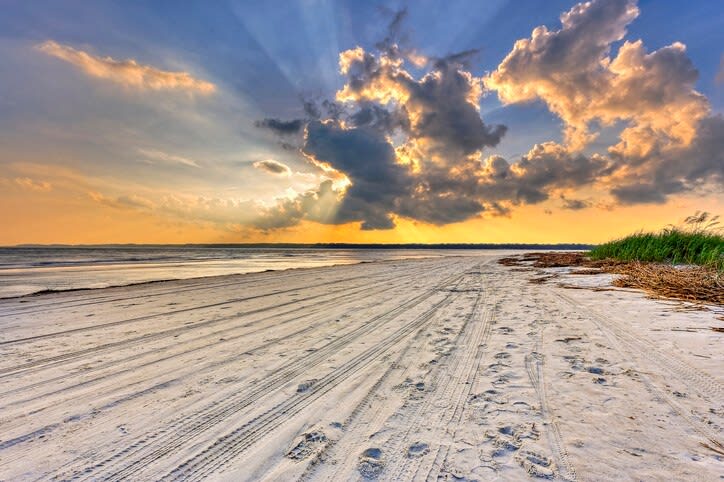 Sunset beach scene: white sandy shore with tire tracks and footprints leading to the ocean, dune grasses on the right, and dramatic clouds with golden sun rays breaking through.