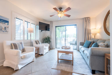 Sunny coastal living room with white slipcovered armchairs, gray sofa with blue pillows, wooden coffee table, ceiling fan, wall-mounted TV, and sliding glass doors opening to a waterfront balcony