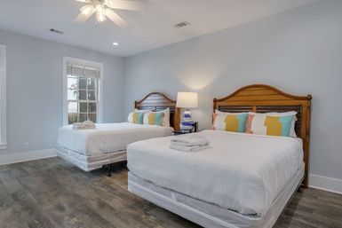 Bright, inviting guest bedroom with two queen beds featuring wooden headboards, crisp white linens, colorful geometric pillows, bedside lamp, ceiling fan, and wide-plank wood-look flooring.