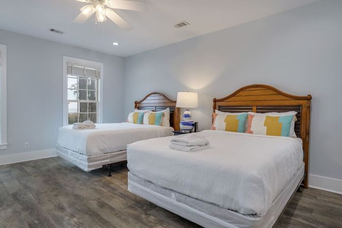 Bright, inviting guest bedroom with two queen beds featuring wooden headboards, crisp white linens, colorful geometric pillows, bedside lamp, ceiling fan, and wide-plank wood-look flooring.