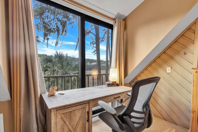 Cozy coastal home office with a wooden desk, ergonomic mesh chair and glowing lamp beside sliding glass doors opening to a balcony overlooking palm trees and a tidal marsh under a bright blue sky.