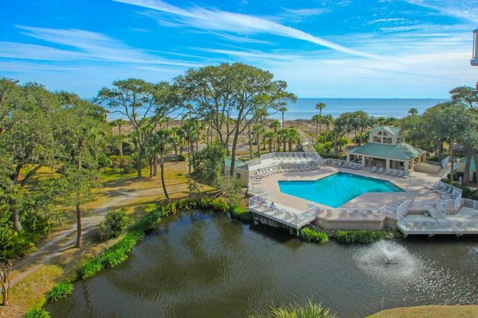 Aerial view of a sunny coastal resort pool with turquoise water, white deck and lounge chairs beside a small lake with a fountain, palm trees and a boardwalk leading to the sandy beach and ocean.
