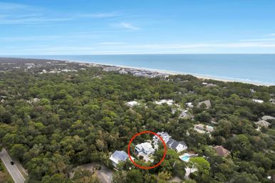 Aerial drone view of a coastal neighborhood with a white house highlighted by a red circle amid dense green tree canopy, nearby sandy beach and calm blue ocean on the horizon.