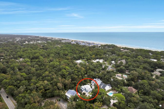 Aerial drone view of a coastal neighborhood with a white house highlighted by a red circle amid dense green tree canopy, nearby sandy beach and calm blue ocean on the horizon.