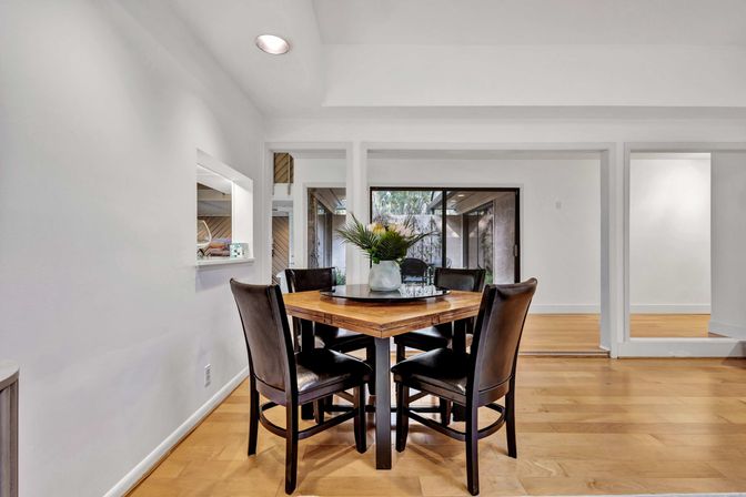 Bright contemporary dining area in a modern home with a square wooden table, four black leather chairs, vase of greenery centerpiece, light hardwood floors, white walls, pass-through kitchen window and sliding glass door to a patio.
