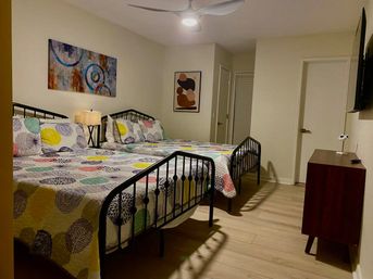 Cozy modern guest bedroom with two black metal-framed double beds dressed in colorful circular-pattern quilts, bedside lamp, abstract wall art, ceiling fan, wall-mounted TV and wooden dresser on light wood floors.