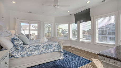 Sunlit coastal bedroom with a white sleigh bed dressed in blue floral bedding, wall of windows showing palm trees and neighboring beach homes, wall-mounted TV and hardwood floors.