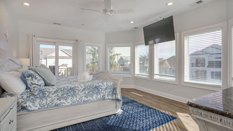 Sunlit coastal bedroom with a white sleigh bed dressed in blue floral bedding, wall of windows showing palm trees and neighboring beach homes, wall-mounted TV and hardwood floors.