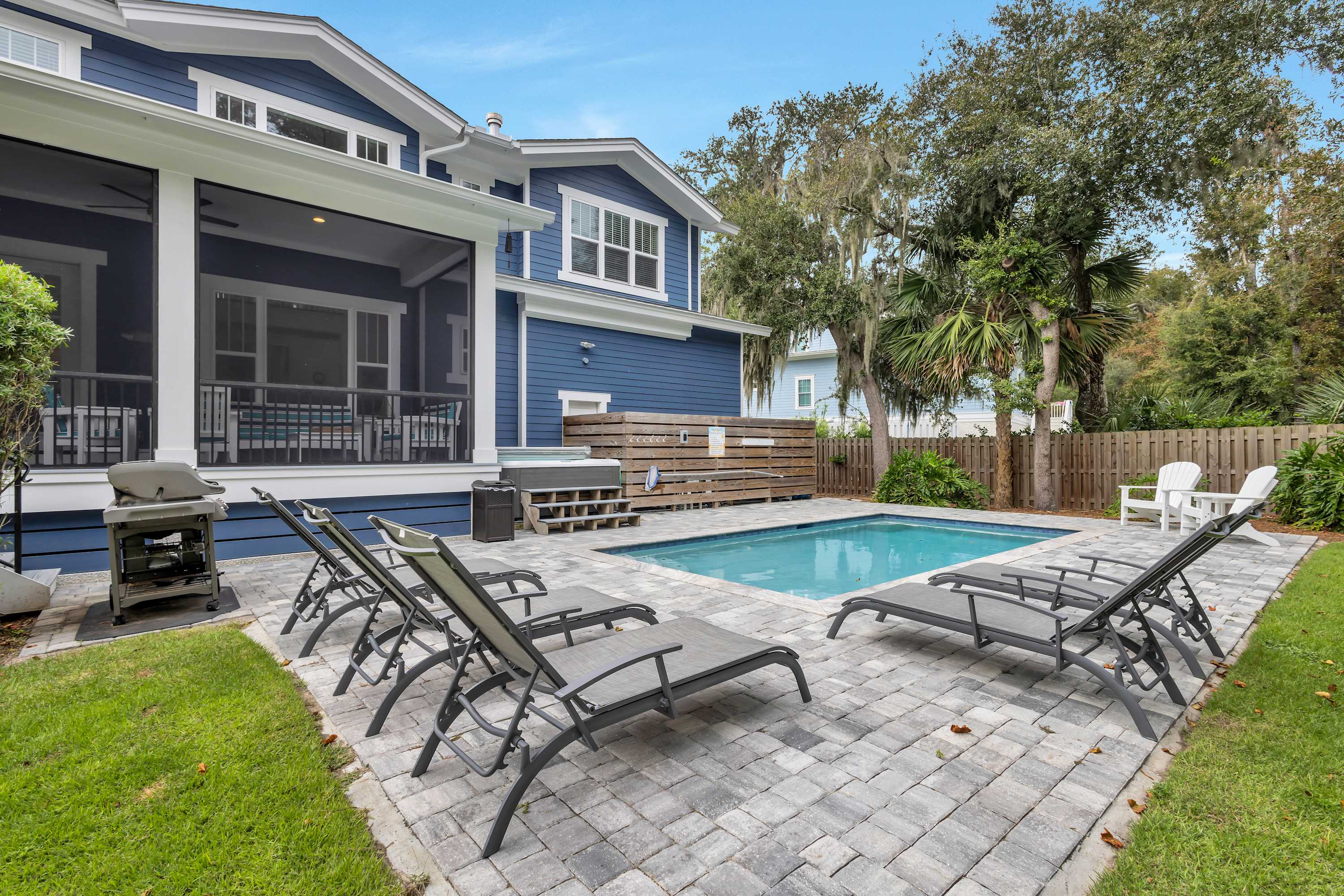 Rectangular backyard pool with gray paver patio, reclining lounge chairs and a grill next to a blue two-story home with a screened porch, wooden privacy fence and palm trees