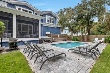 Rectangular backyard pool with gray paver patio, reclining lounge chairs and a grill next to a blue two-story home with a screened porch, wooden privacy fence and palm trees