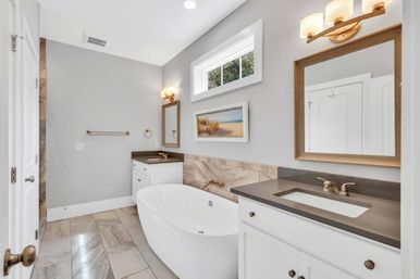 Spa-like modern bathroom with a white freestanding soaking tub, gray quartz vanity with undermount sink, wood-framed mirror and brass fixtures, marble-look floor tile and a small high window