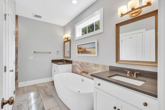 Spa-like modern bathroom with a white freestanding soaking tub, gray quartz vanity with undermount sink, wood-framed mirror and brass fixtures, marble-look floor tile and a small high window