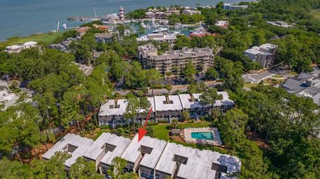 Aerial view of waterfront townhomes and marina with a lighthouse; red arrow marks a rooftop unit amid pine trees and a resort-style pool.