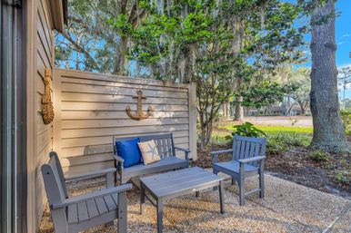Cozy coastal-style patio with gray wooden bench, two chairs and coffee table, blue cushions and starfish pillow, rope anchor wall decor on shiplap fence, gravel floor, shaded by oak trees draped in Spanish moss.