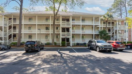 Sunlit three-story beige condominium with white exterior walkways and railings, shaded parking lot with parked SUVs, pine and palm trees under a blue sky.