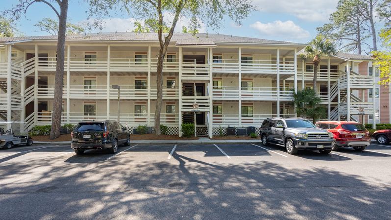 Sunlit three-story beige condominium with white exterior walkways and railings, shaded parking lot with parked SUVs, pine and palm trees under a blue sky.