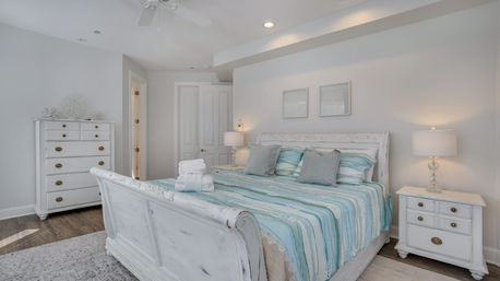 Bright coastal-style master bedroom with white distressed sleigh bed, turquoise-striped bedding and gray accent pillows, matching white nightstands and dresser, glass table lamps, hardwood floors and folded towels on the bed.