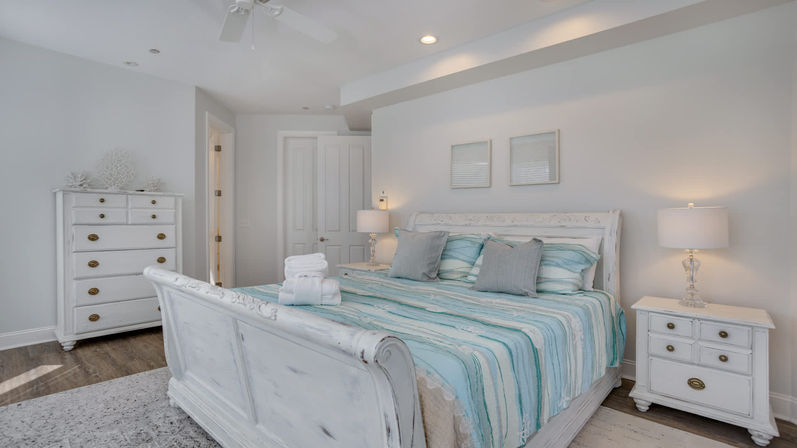 Bright coastal-style master bedroom with white distressed sleigh bed, turquoise-striped bedding and gray accent pillows, matching white nightstands and dresser, glass table lamps, hardwood floors and folded towels on the bed.