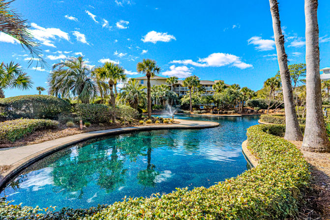 Sunny tropical coastal resort lagoon with palm trees, a central fountain, reflective blue water, landscaped walking path and low-rise vacation condos